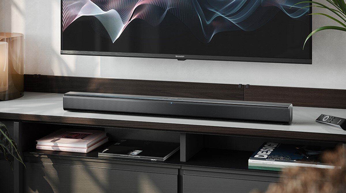 Na obrazie znajduje się czarny soundbar umieszczony na półce pod telewizorem. W tle widoczny jest fragment wnętrza z rośliną i kilkoma książkami na półce.