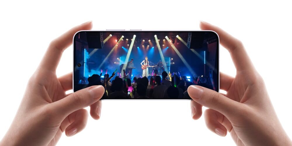 Osoba trzyma smartfon, na ekranie którego widać koncert z kolorowymi światłami i muzykami na scenie. Tło jest białe, a dłonie są jedynym widocznym elementem poza urządzeniem.
