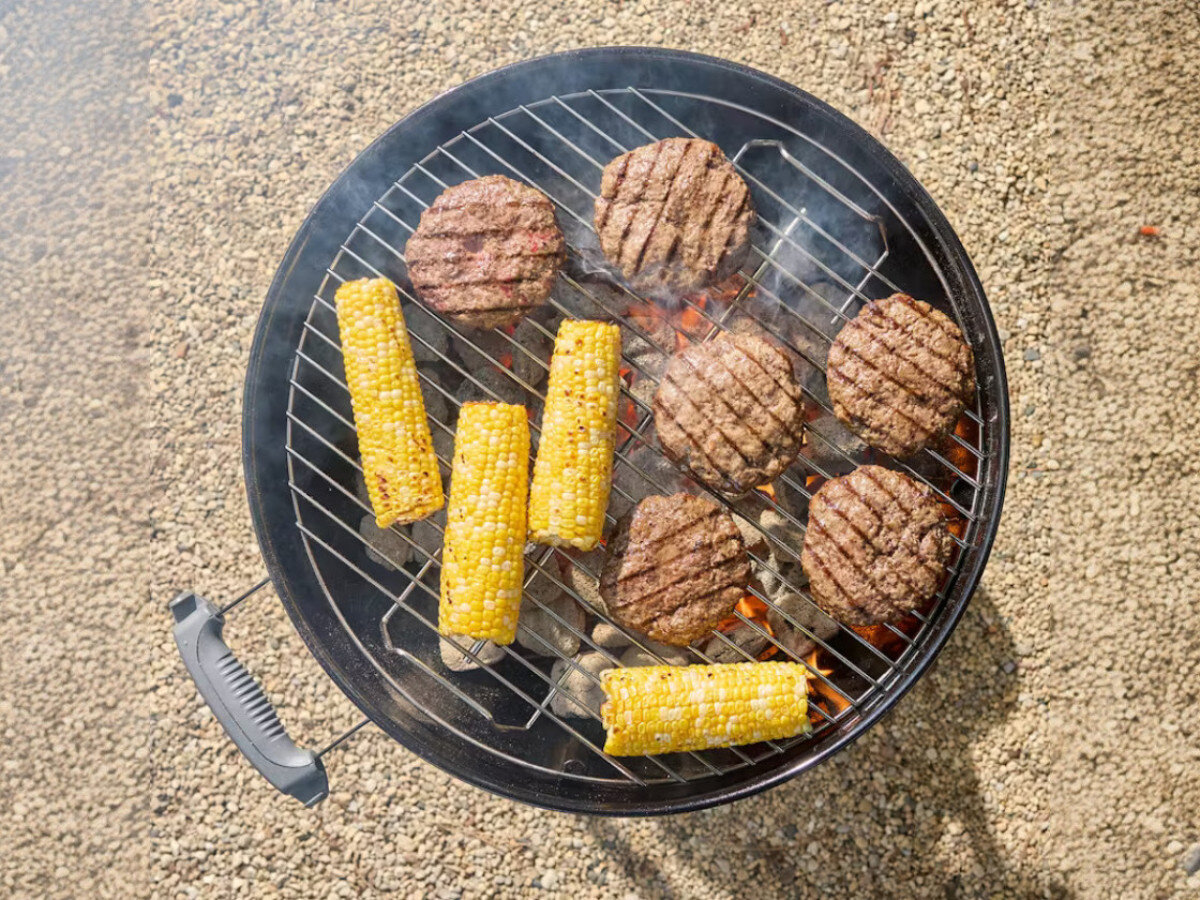 Na okrągłym grillu węglowym znajdują się grillowane burgery i kolby kukurydzy. Grill stoi na żwirowym podłożu, a z wnętrza unosi się dym.