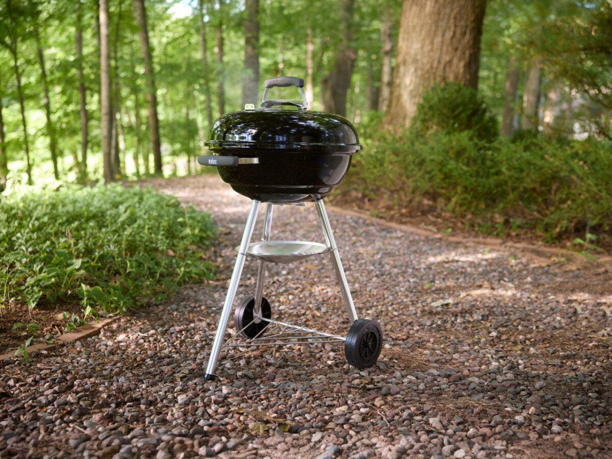 Czarny, okrągły grill węglowy na kółkach stoi na żwirowej ścieżce w lesie. Grill ma pokrywę z uchwytem i metalowe nogi.