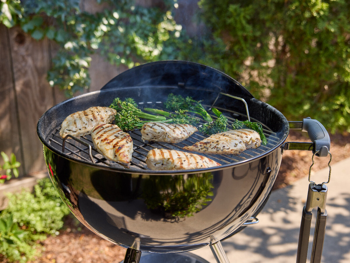 Na okrągłym grillu węglowym znajdują się grillowane kawałki mięsa i zielone warzywa. W tle widoczne są rośliny i drewniany płot.