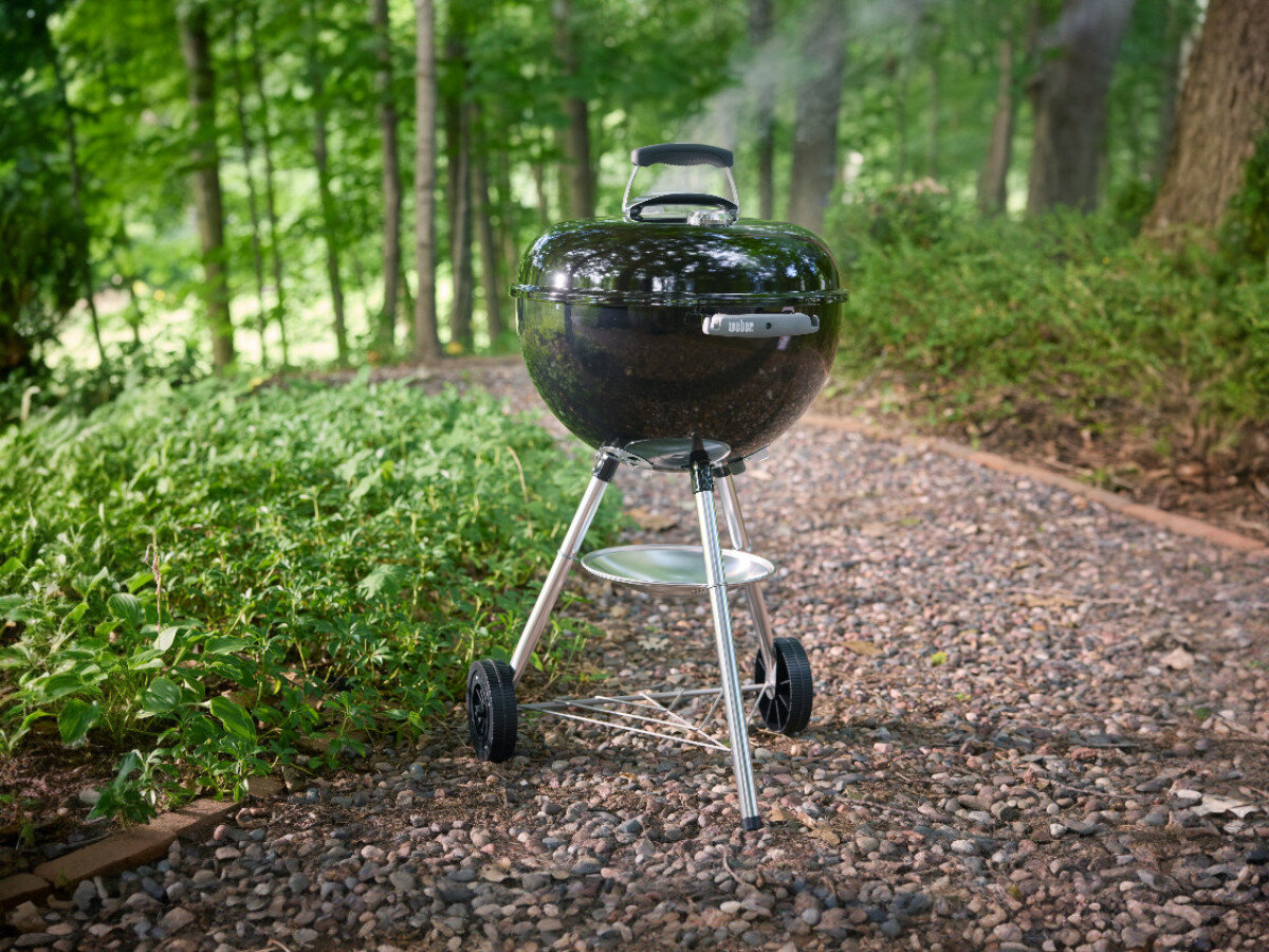 Okrągły grill węglowy na kółkach stoi na żwirowej ścieżce w otoczeniu zieleni. Na pokrywie widoczne jest logo \'Weber\'.
