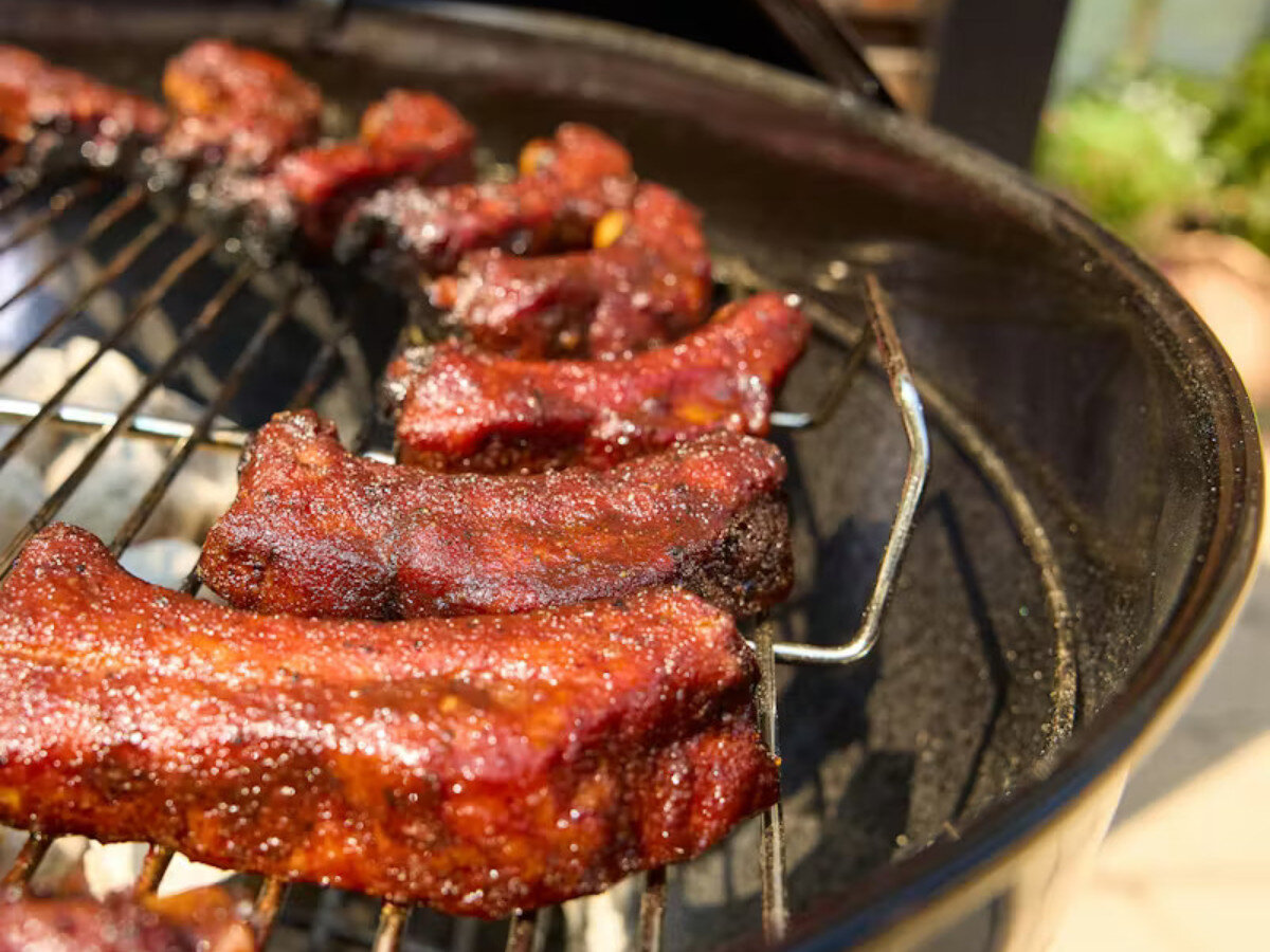 Na grillu węglowym znajdują się kawałki mięsa z widoczną przypieczoną skórką. Grill jest okrągły i posiada metalowy ruszt.