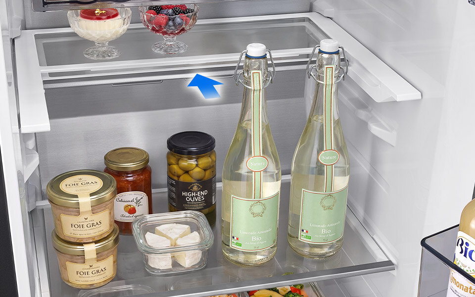 składana półka Wnętrze lodówki z widocznymi półkami, na których znajdują się różne produkty spożywcze, takie jak słoiki z foie gras, oliwki, butelki z napojami oraz deser w pucharku. Na jednej z półek widoczna jest niebieska strzałka wskazująca na przestrzeń pomiędzy półkami.