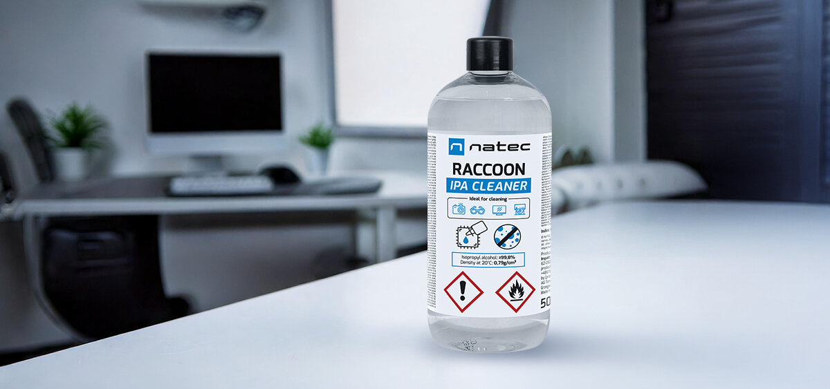 Na obrazie widoczna jest butelka z etykietą \'natec Raccoon IPA Cleaner\' o pojemności 500 ml, stojąca na białym stole w nowoczesnym wnętrzu biurowym. Etykieta zawiera symbole ostrzegawcze oraz informacje o zastosowaniu produktu.