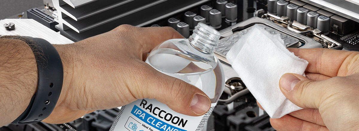 Osoba trzyma butelkę z napisem \'RACCOON IPA CLEANER\' i wylewa płyn na biały materiał. W tle widoczny jest fragment wnętrza komputera z elementami elektronicznymi.
