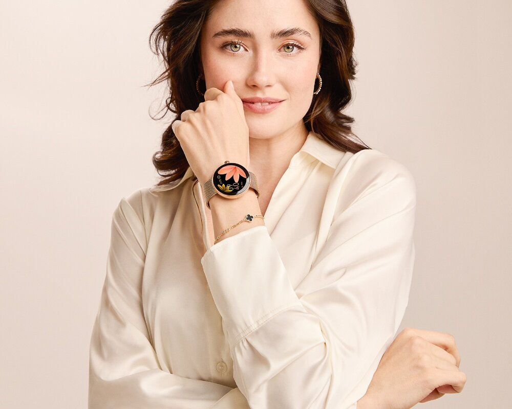 Osoba w białej koszuli prezentuje smartwatch z czarnym wyświetlaczem, na którym widoczny jest motyw kwiatowy. Na nadgarstku znajduje się również delikatna bransoletka.