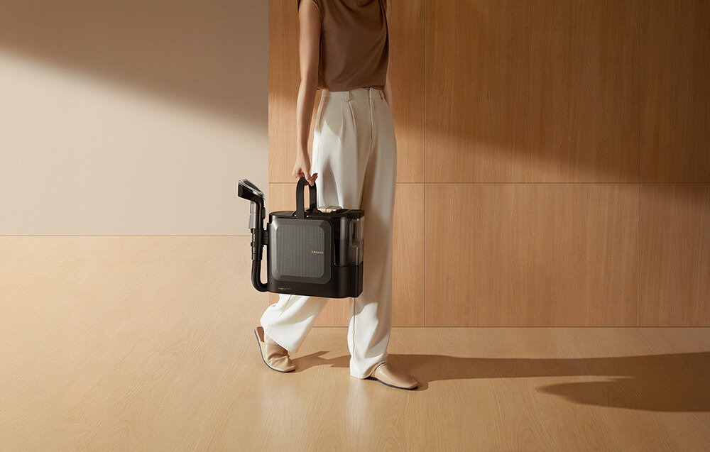 Osoba trzyma w ręce odkurzacz piorący DREAME N20 Steam w minimalistycznym wnętrzu z drewnianą podłogą i ścianą. Odkurzacz ma ciemny kolor i kompaktowy kształt.