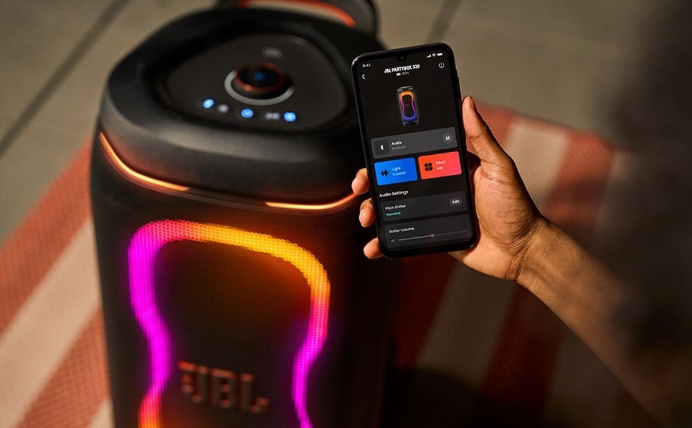Osoba trzyma smartfon z aplikacją kontrolującą głośnik JBL PartyBox 330, który emituje kolorowe światła. Na głośniku widoczne jest logo \'JBL\'.