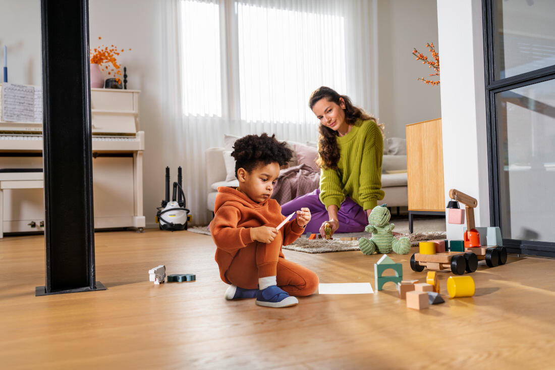W jasnym pokoju dziecko w pomarańczowym ubraniu bawi się na podłodze, a w tle dorosła osoba siedzi na dywanie. W rogu pomieszczenia stoi parownica KARCHER SC 3 EasyFix.