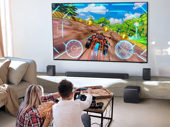 Dwie osoby grające w wyścigi na telewizorze z soundbarem Samsung HW-Q990H i głośnikami ustawionymi wokół ekranu.