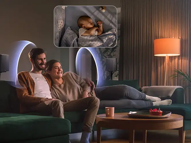 Para siedząca na kanapie w salonie z zapaloną lampą, głośnikiem Samsung i niebieskimi kręgami dźwięku za plecami.
