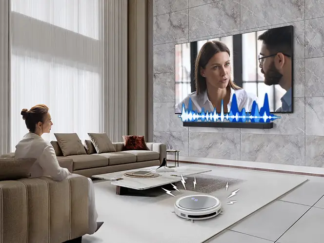 Kobieta ogląda rozmowę na telewizorze z soundbarem Samsung HW-Q930H, obok robota sprzątającego i niebieskich fal dźwięku.