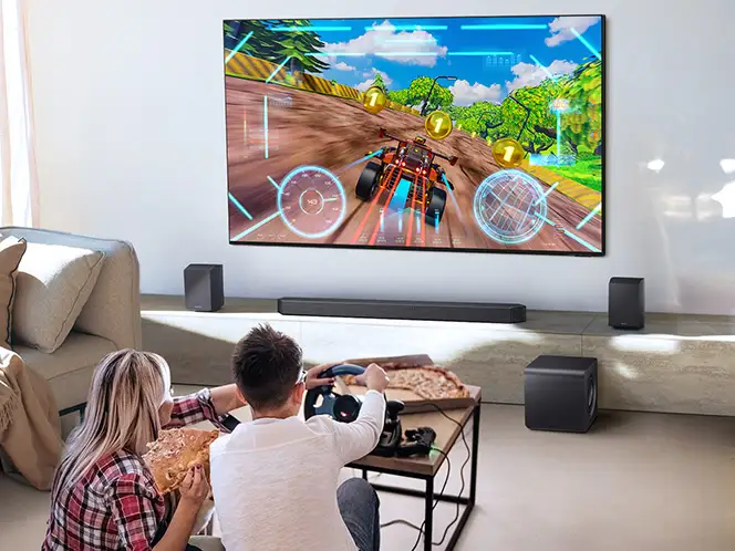 Dwie osoby grające w wyścigi na telewizorze z soundbarem Samsung HW-Q930H i głośnikami ustawionymi wokół ekranu.
