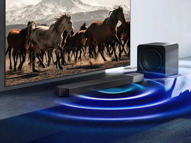 Telewizor z biegnącymi końmi, soundbar Samsung HW-Q930H i subwoofer z niebieskimi falami dźwięku na podłodze.