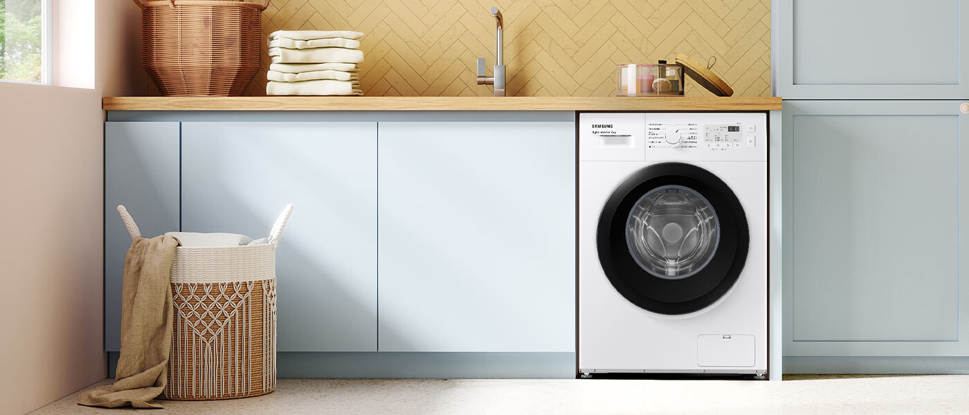 W kuchni znajduje się biała pralka w zabudowie z widocznym panelem sterowania i okrągłym oknem bębna. Obok pralki stoi kosz na pranie z ręcznikiem, a na blacie leżą złożone ręczniki i kosz wiklinowy.