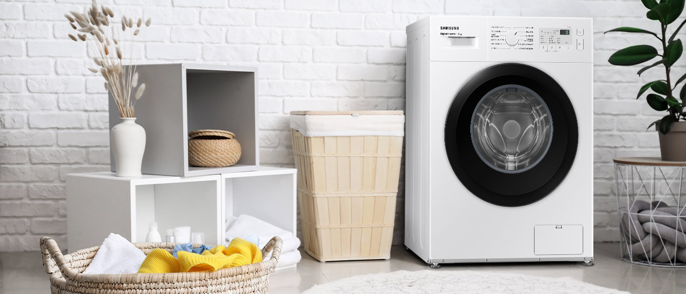 Na obrazie znajduje się biała pralka z widocznym panelem sterowania i logo \'SAMSUNG\'. Obok pralki stoi kosz na pranie, a w tle widać białą ceglaną ścianę oraz dekoracyjne półki z przedmiotami.