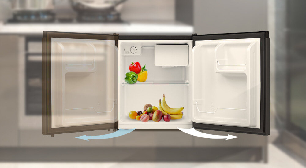 Otwarta lodówka z widocznymi owocami i warzywami na półce, w tym banany, jabłka, papryka i pomidory. W tle rozmyta kuchnia z widocznymi garnkami na kuchence.