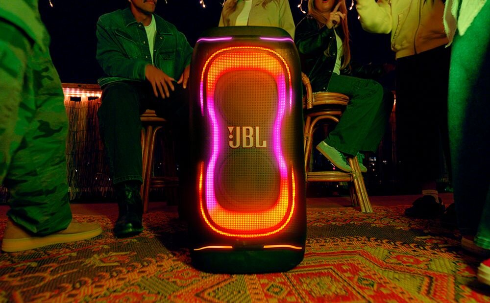 Na obrazie widać czarny głośnik z podświetleniem LED w kolorach czerwonym i fioletowym, z logo \'JBL\' na środku. W tle znajdują się osoby siedzące na krzesłach oraz dywan na podłodze.