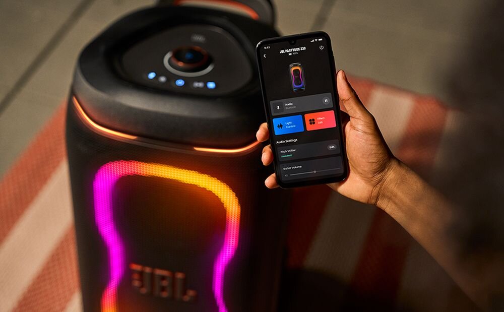 Osoba trzyma smartfon z aplikacją kontrolującą głośnik JBL PartyBox 330, który emituje kolorowe światła. Na głośniku widoczne jest logo \'JBL\'.