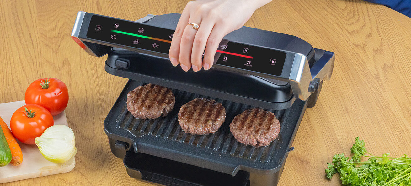Osoba otwiera grill elektryczny, na którym znajdują się trzy grillowane kotlety. Obok grilla leży deska z pokrojonymi warzywami i ziołami.