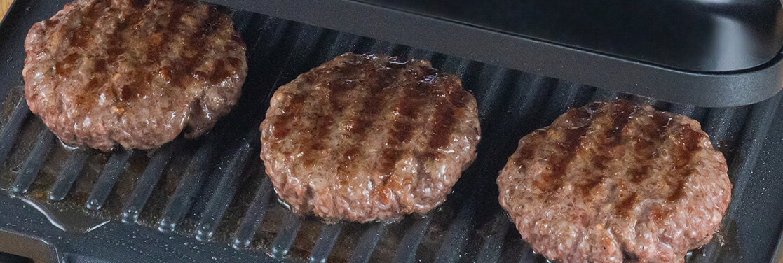 Trzy burgery grillują się na elektrycznym grillu z widocznymi śladami rusztu. Grill ma czarną powierzchnię z rowkami do odprowadzania tłuszczu.
