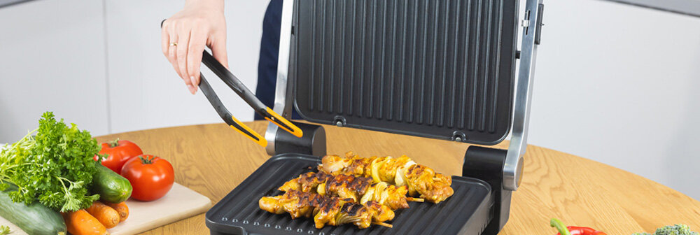 Osoba używa szczypiec do obracania grillowanego mięsa na elektrycznym grillu z otwartą pokrywą. Obok na stole znajdują się świeże warzywa, takie jak pomidory i sałata.