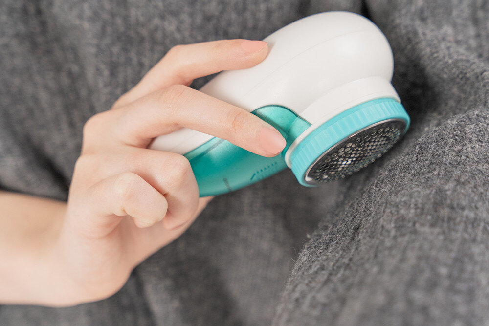Osoba używa białej i turkusowej golarki do ubrań na szarej tkaninie. Golarka ma okrągłą, perforowaną powierzchnię roboczą.