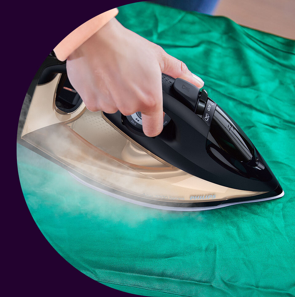 Osoba prasuje zielony materiał za pomocą czarnego żelazka, z którego wydobywa się para. Na żelazku widoczne są przyciski i pokrętło, a na stopie napis \'PHILIPS\'.