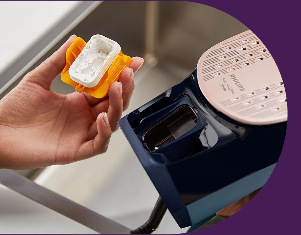 Osoba trzyma żółty pojemnik z białą substancją, obok znajduje się żelazko z widoczną stopą i napisem \'PHILIPS SteamGlide Elite\'.