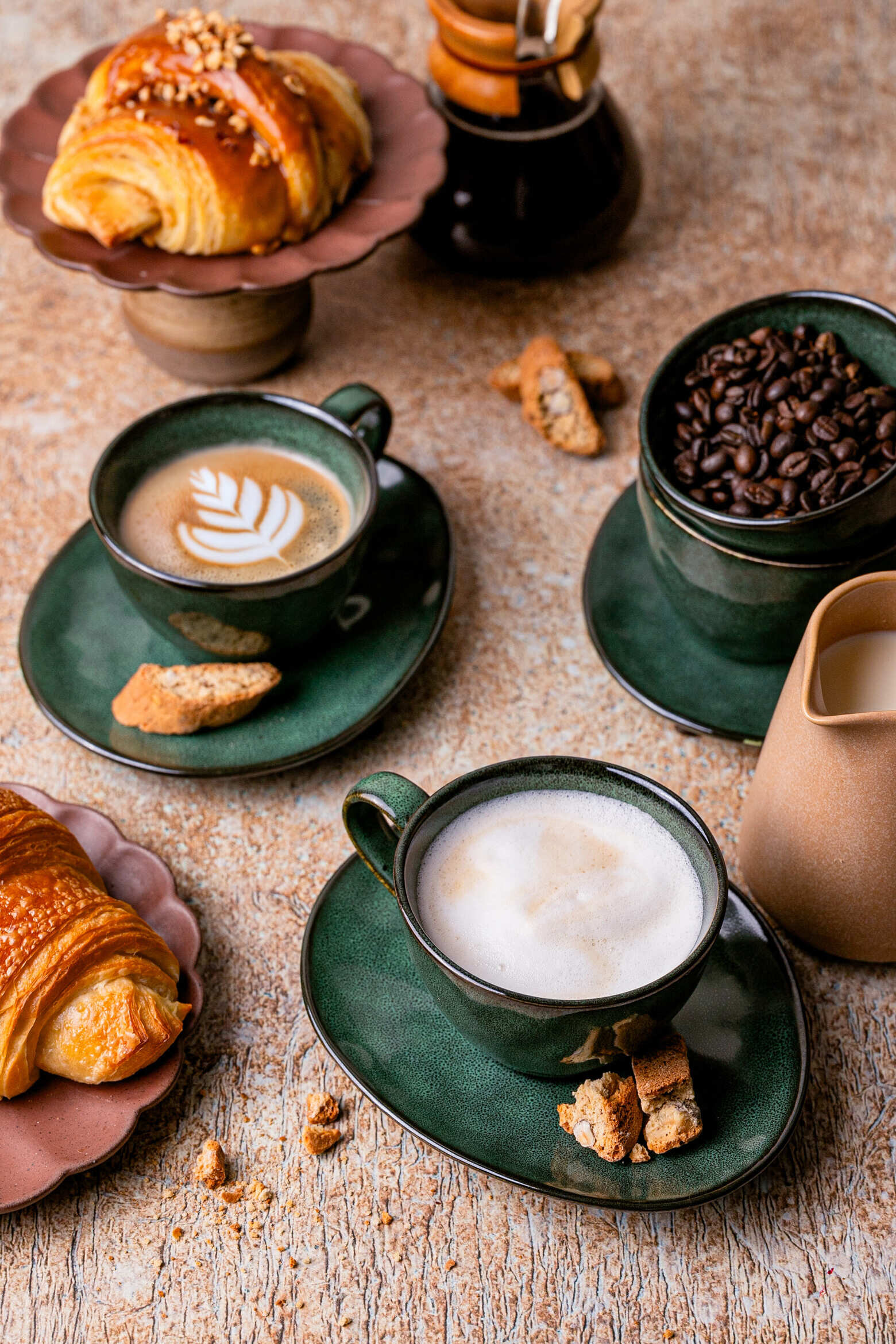 Na obrazie widoczne są zielone filiżanki z kawą na spodkach, obok których leżą kawałki ciasta i croissant. W tle znajduje się dzbanek z mlekiem oraz filiżanka z ziarnami kawy.