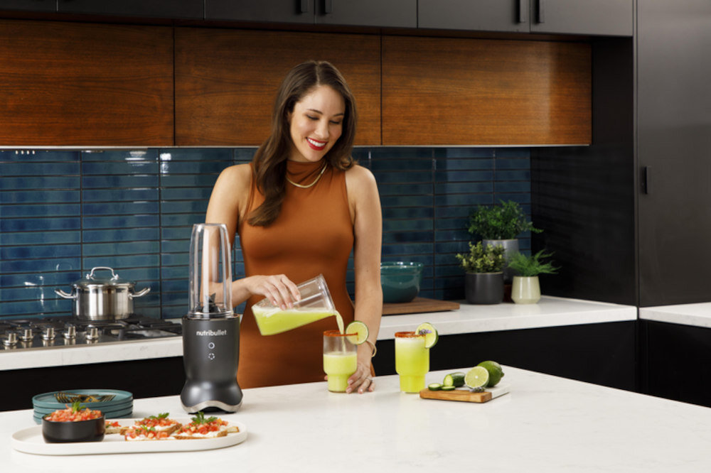 Kobieta w kuchni nalewa zielony napój do szklanki, obok stoi blender z logo \'nutribullet\'. Na blacie znajdują się pokrojone limonki i inne składniki.