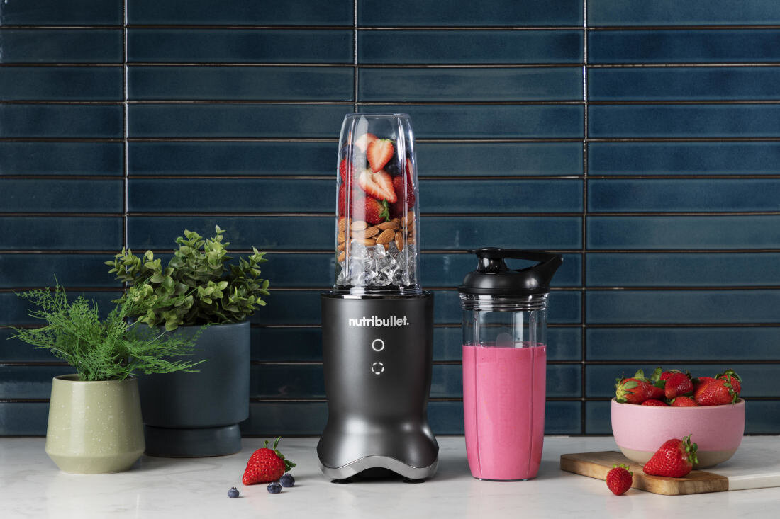 Na blacie kuchennym stoi blender personalny z logo \'nutribullet\', wypełniony truskawkami, migdałami i lodem. Obok znajduje się kubek z różowym koktajlem oraz miska z truskawkami, a w tle widoczne są doniczki z roślinami.
