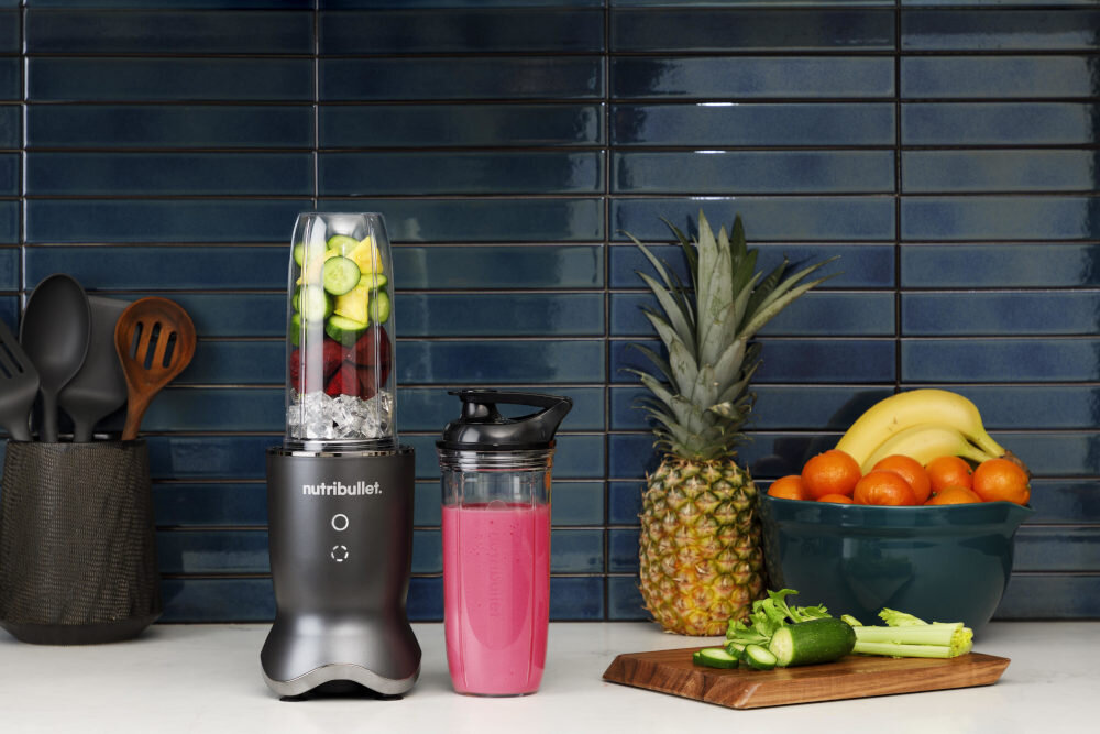 Na blacie kuchennym stoi blender z logo \'nutribullet\', wypełniony kawałkami owoców i lodem. Obok znajduje się pojemnik z różowym napojem, a w tle widoczne są owoce i przybory kuchenne.