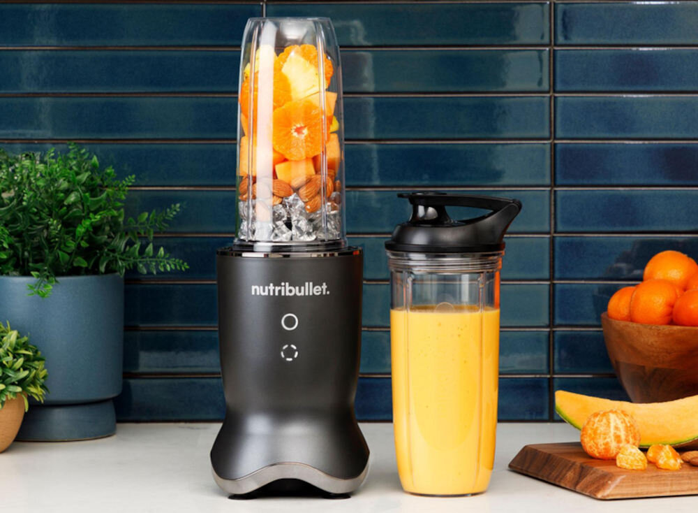 Na blacie kuchennym stoi blender z logo \'nutribullet\', wypełniony kawałkami owoców i lodem. Obok znajduje się zamknięty pojemnik z pomarańczowym napojem, a w tle widoczne są rośliny i miska z owocami.