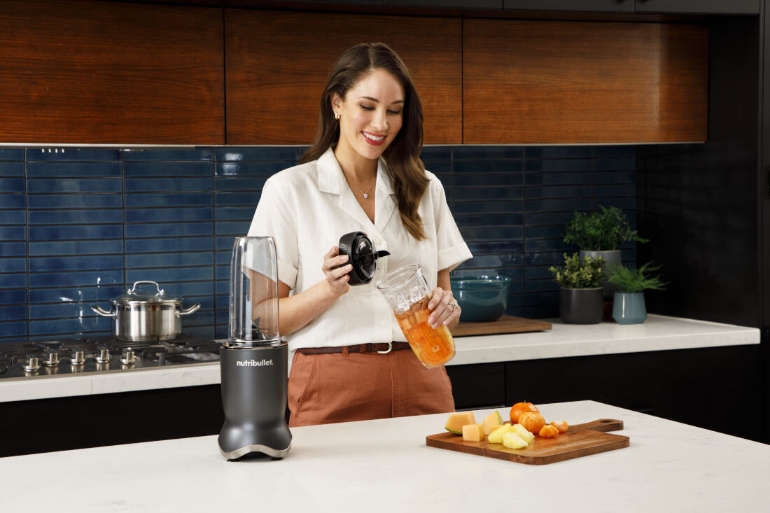 Kobieta w kuchni przygotowuje napój, używając blendera personalnego z logo \'nutribullet\'. Na blacie znajduje się deska z pokrojonymi owocami.