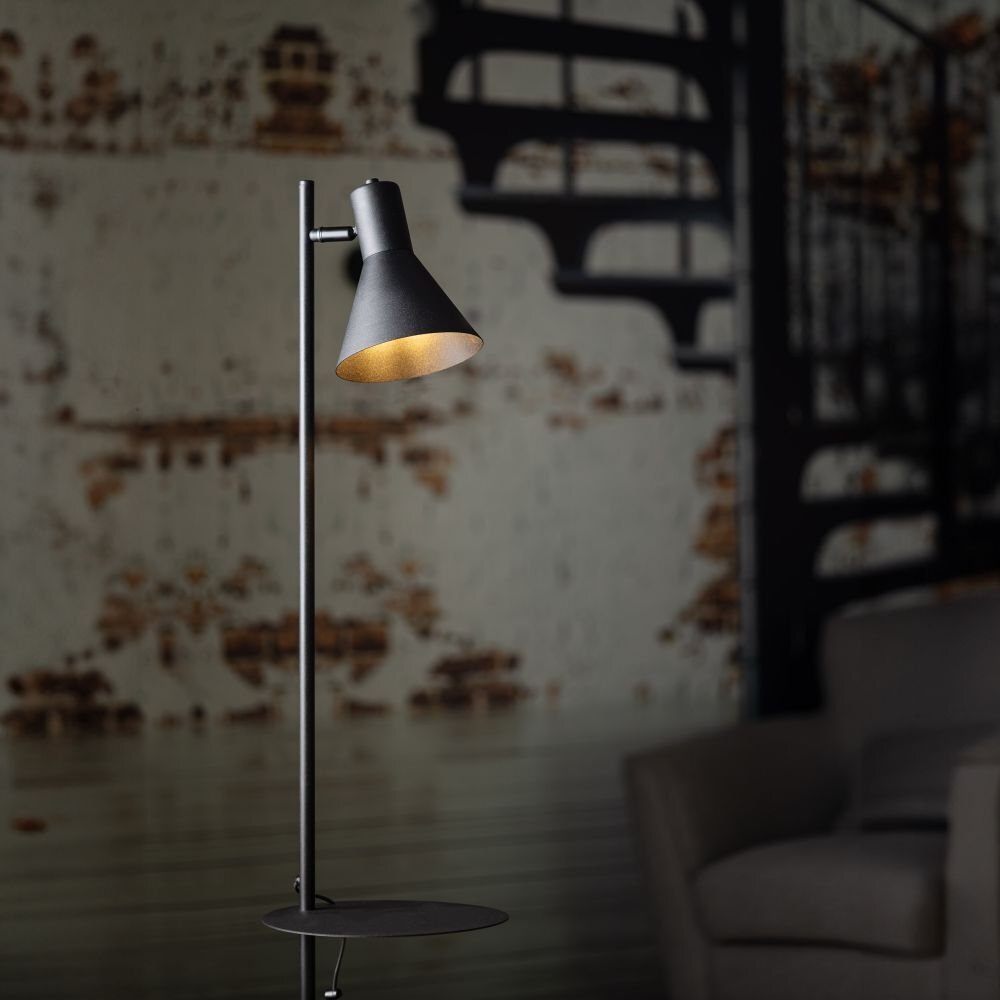 Czarna lampa podłogowa z półką i regulowanym kloszem, ustawiona na tle ściany z widocznymi elementami architektonicznymi.