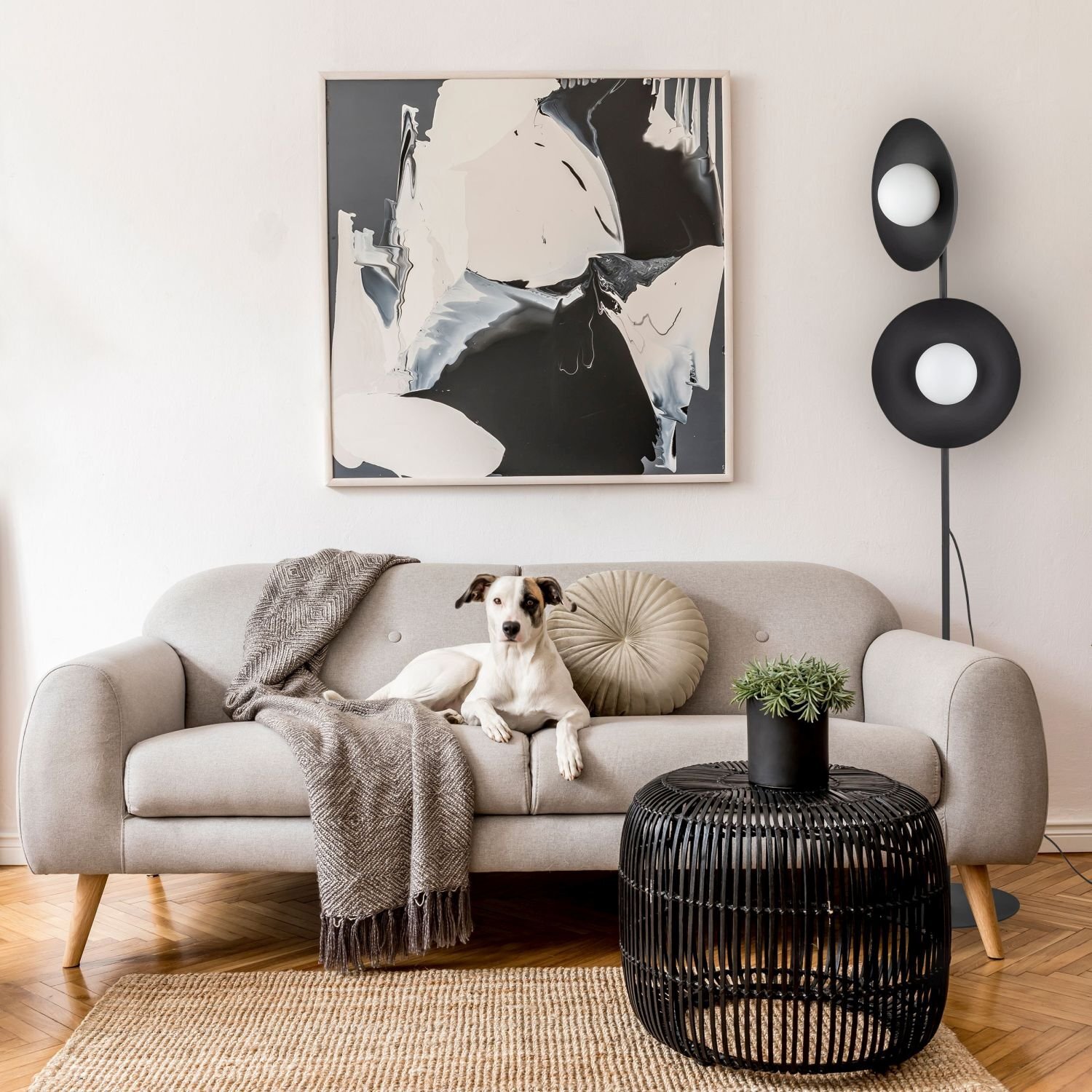 W salonie znajduje się szara sofa z poduszkami i kocem, na której siedzi pies. Obok stoi nowoczesna lampa podłogowa z czarnymi elementami i szklanymi kloszami, a na ścianie wisi abstrakcyjny obraz.
