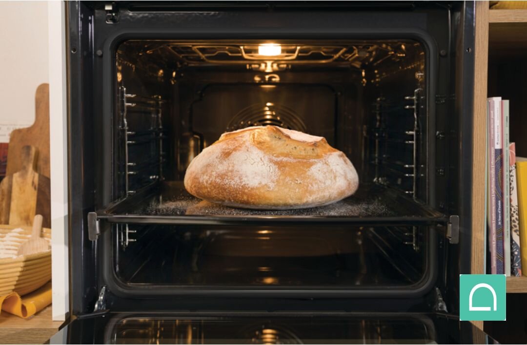 Otwarty piekarnik z oświetleniem wewnętrznym, w którym na środkowej półce znajduje się upieczony bochenek chleba. Obok piekarnika widoczne są książki kucharskie i przybory kuchenne.