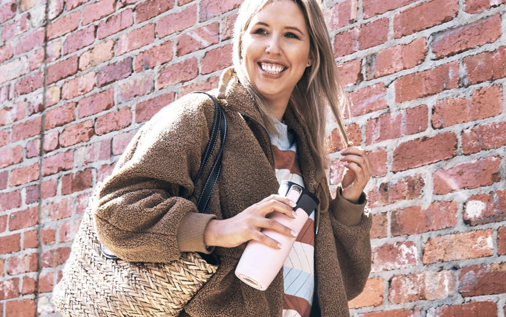 Osoba trzyma różowy kubek termiczny przed ceglanym murem, ubrana w brązowy płaszcz i trzymająca plecioną torbę.