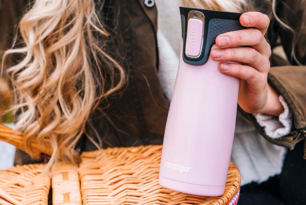 Osoba trzyma różowy kubek termiczny z logo \'contigo\' na tle wiklinowego kosza. Kubek ma czarną pokrywkę z przyciskiem oznaczonym \'AUTOSEAL\'.