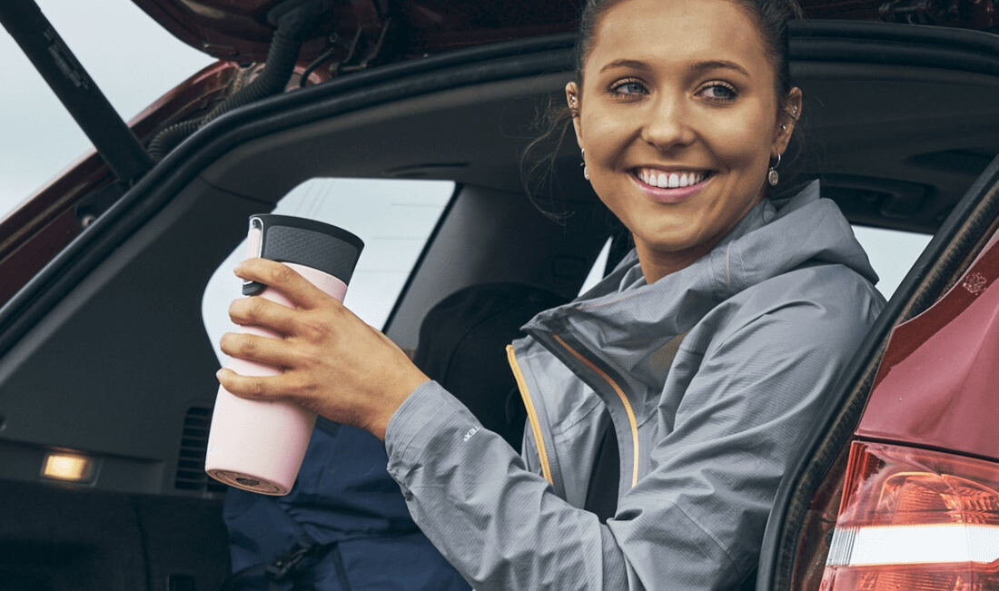 Osoba siedzi w otwartym bagażniku samochodu, trzymając różowy kubek termiczny z czarną pokrywką. W tle widoczny jest plecak.