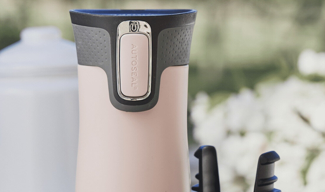 Różowy kubek termiczny z czarną pokrywką i przyciskiem z napisem \'AUTOSEAL\'. W tle widoczne są rozmyte elementy krajobrazu.