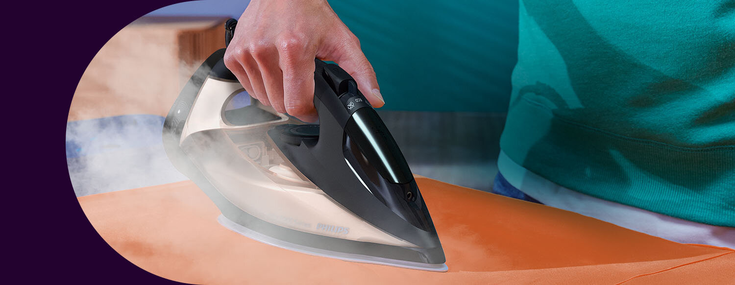 Osoba prasuje pomarańczowy materiał żelazkiem, z którego wydobywa się para. Na żelazku widoczne jest logo \'PHILIPS\'.