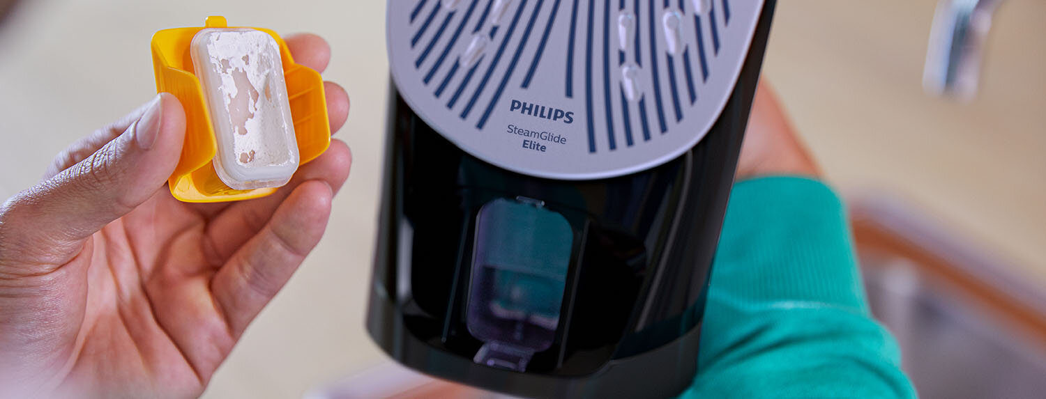 Osoba trzyma żółty pojemnik z białym elementem, obok widoczne jest żelazko z napisem \'PHILIPS SteamGlide Elite\'.