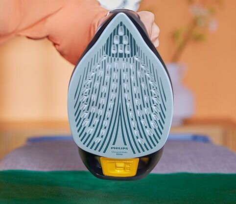 Zdjęcie przedstawia stopę żelazka z widocznymi otworami parowymi i napisem \'PHILIPS Azur\'. W tle rozmyty widok wnętrza z kwiatami w wazonie.