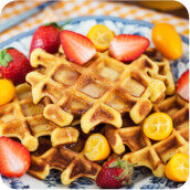 Na talerzu znajdują się gofry udekorowane truskawkami i kawałkami pomarańczowych owoców. Tło to kraciasty obrus w kolorze pomarańczowo-białym.