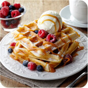 Na talerzu znajdują się gofry z gałką lodów waniliowych, polane sosem karmelowym i udekorowane malinami oraz jagodami. Obok stoi filiżanka z napojem i miseczka z malinami.