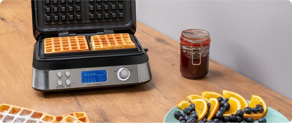 Gofrownica z dwoma goframi na drewnianym blacie, obok słoik z ciemnym płynem i talerz z owocami. Na wyświetlaczu widoczny jest czas 08:55, a poniżej napis GÖTZE & JENSEN.