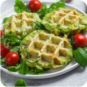 Na talerzu znajdują się trzy zielone gofry otoczone liśćmi sałaty i pomidorkami koktajlowymi. Tło jest jasne i neutralne.